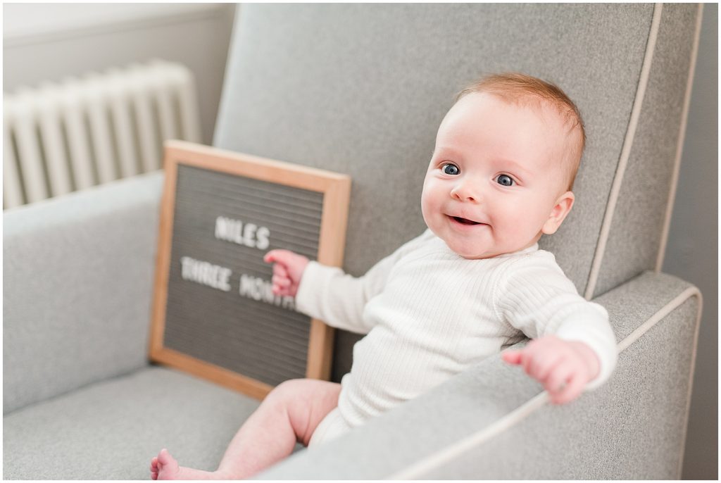 Richmond photographers niles laughing on grey chair with letter board at 3 months