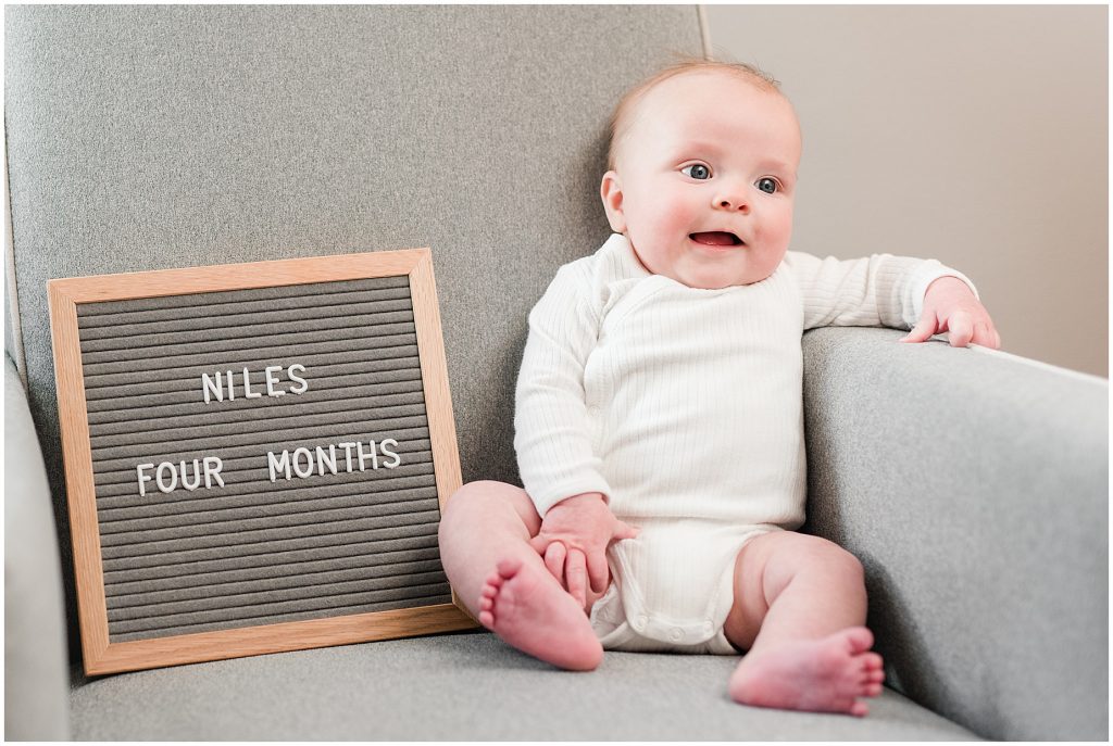 Richmond photographers niles sitting on grey chair with letter board at 4 months