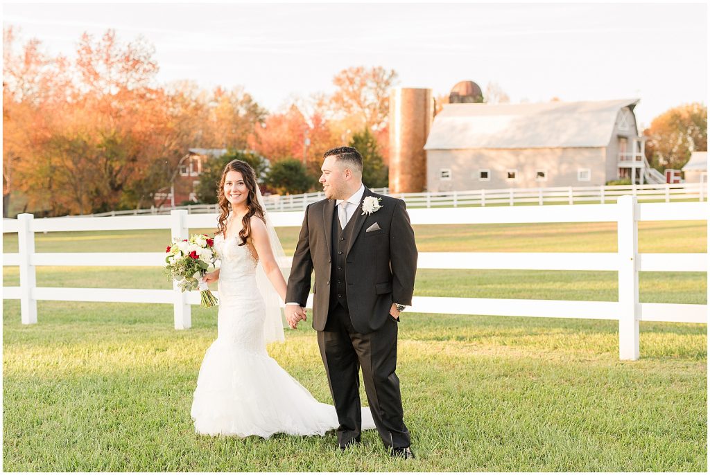 bride and groom walking in field with barn and fall trees behind them at amber grove