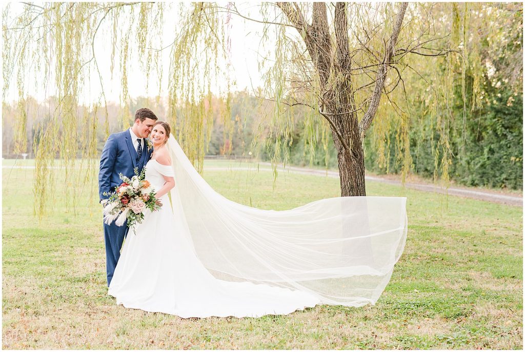 bride's veil flowing in the wind with groom standing in a willow tree at Waverly Estate in southern Virginia